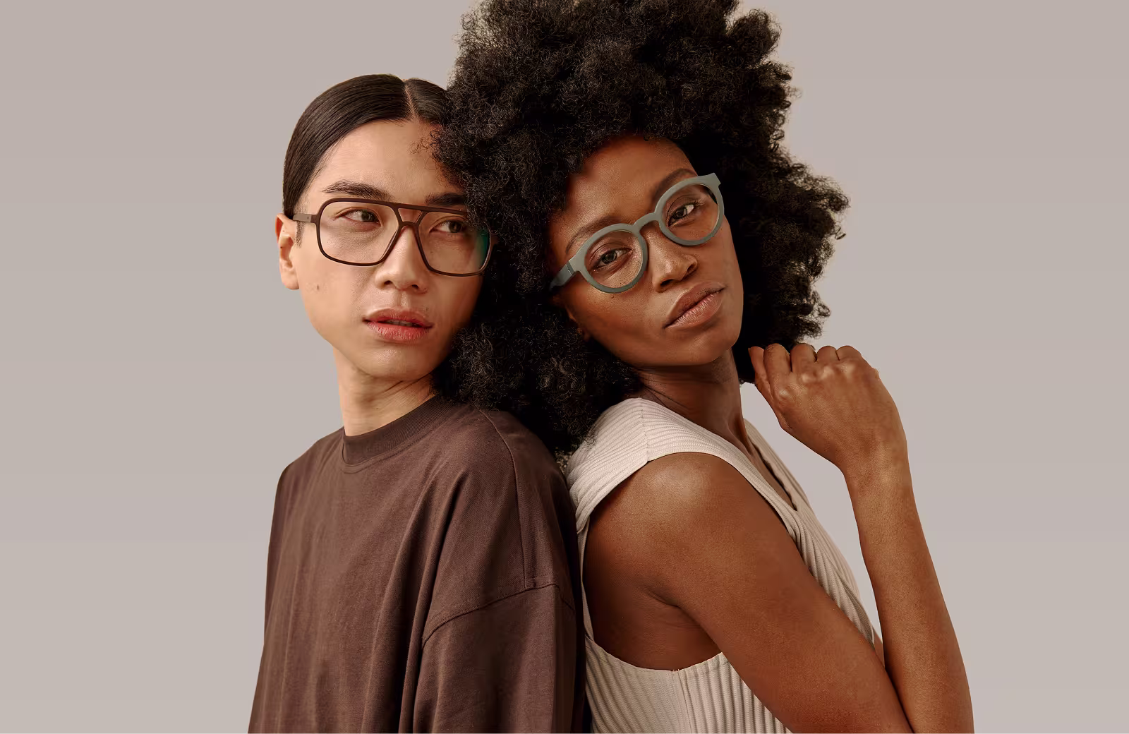 Ein Mann und eine Frau mit stilvollen Brillen lehnen Rücken an Rücken vor einem neutralen Hintergrund.