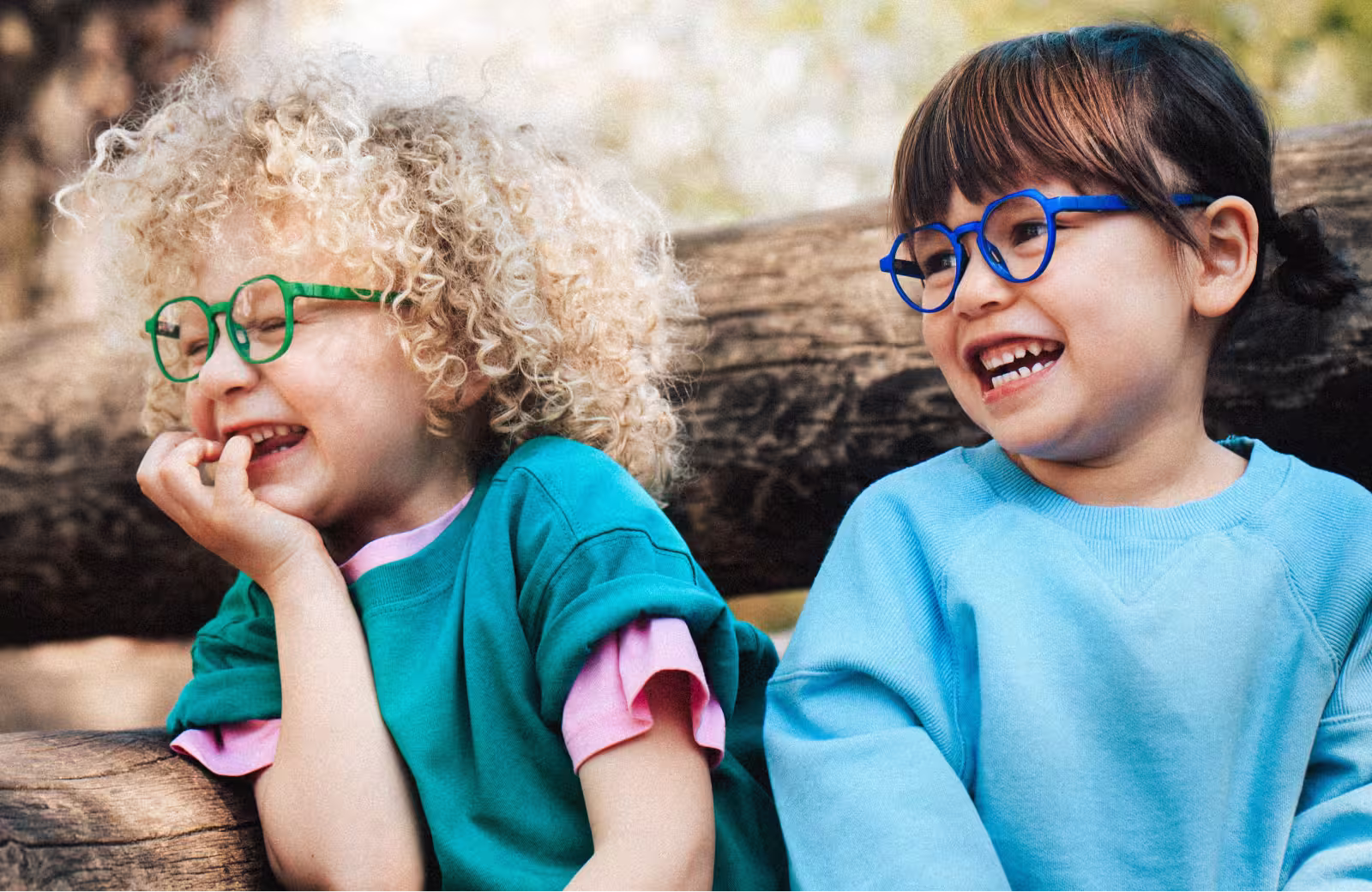 Zwei Kinder mit bunten Brillen lächeln und sitzen auf einem Holzstamm im Freien.