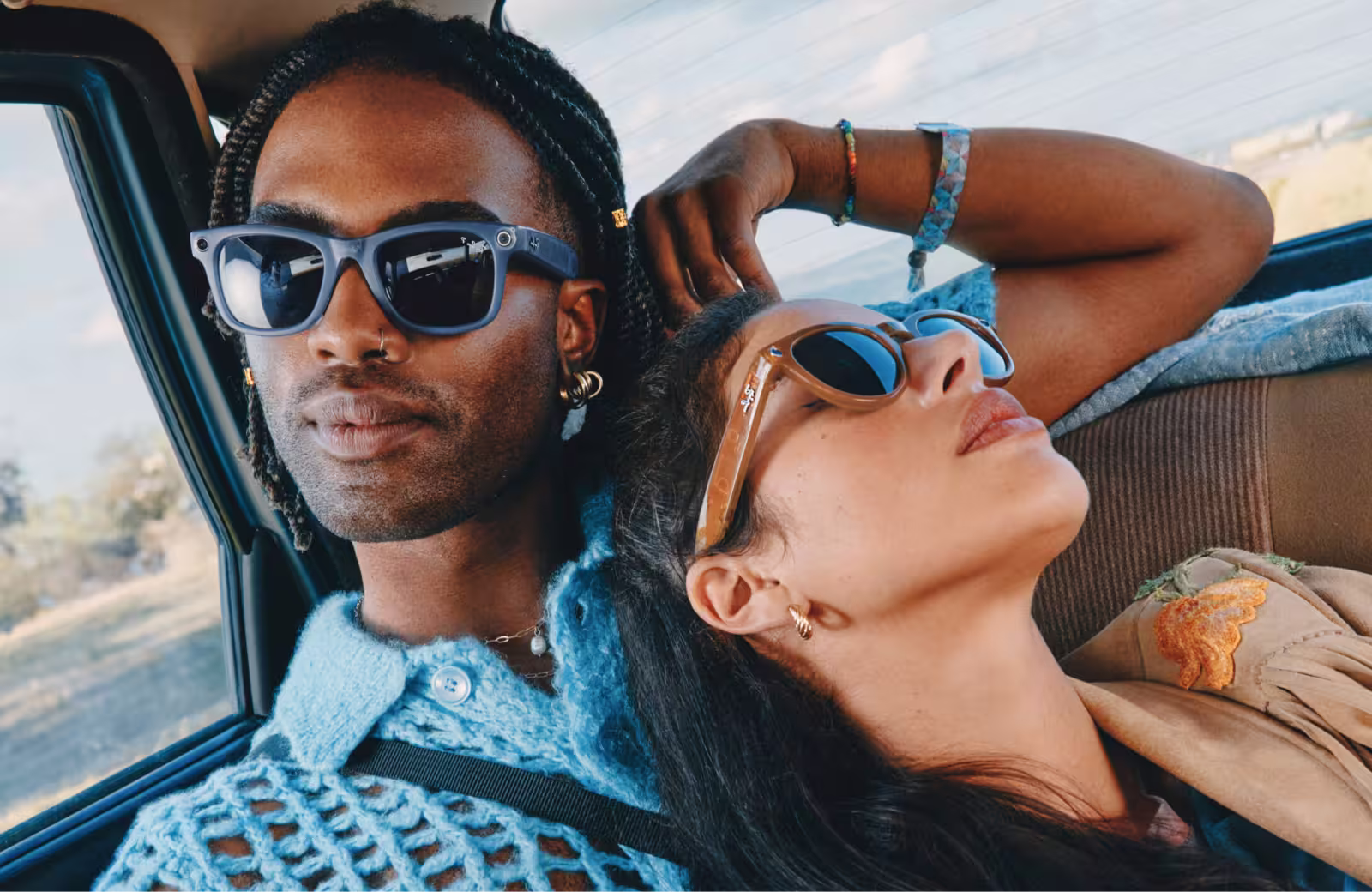 Zwei Personen mit Sonnenbrille sitzen eng beieinander in einem Fahrzeug, eine lehnt sich zurück und stützt den Arm hinter dem Kopf ab.