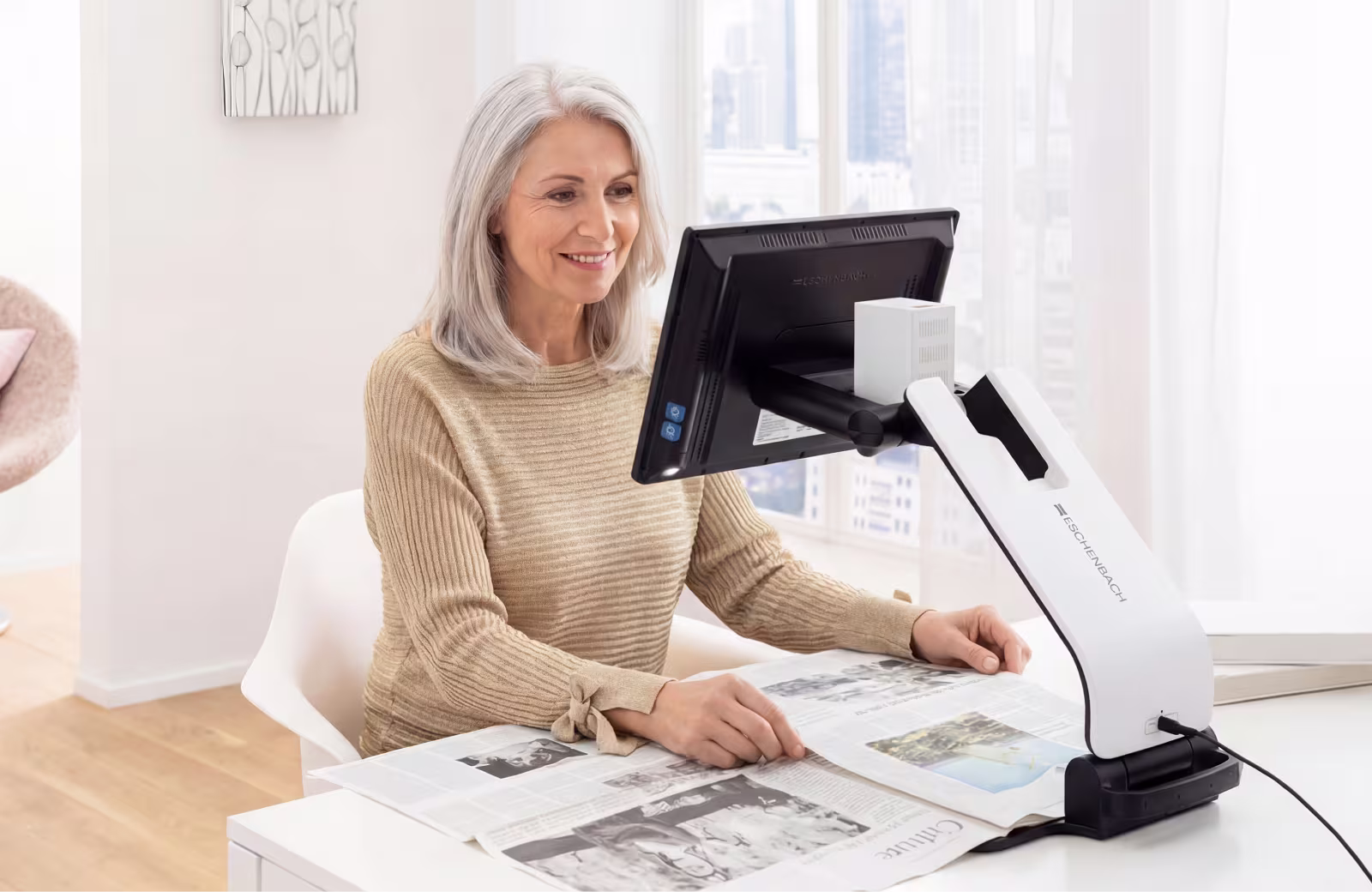 Lächelnde ältere Frau, die ein elektronisches Eschenbach-Lesegerät benutzt, um eine Zeitung an einem weißen Schreibtisch zu lesen.